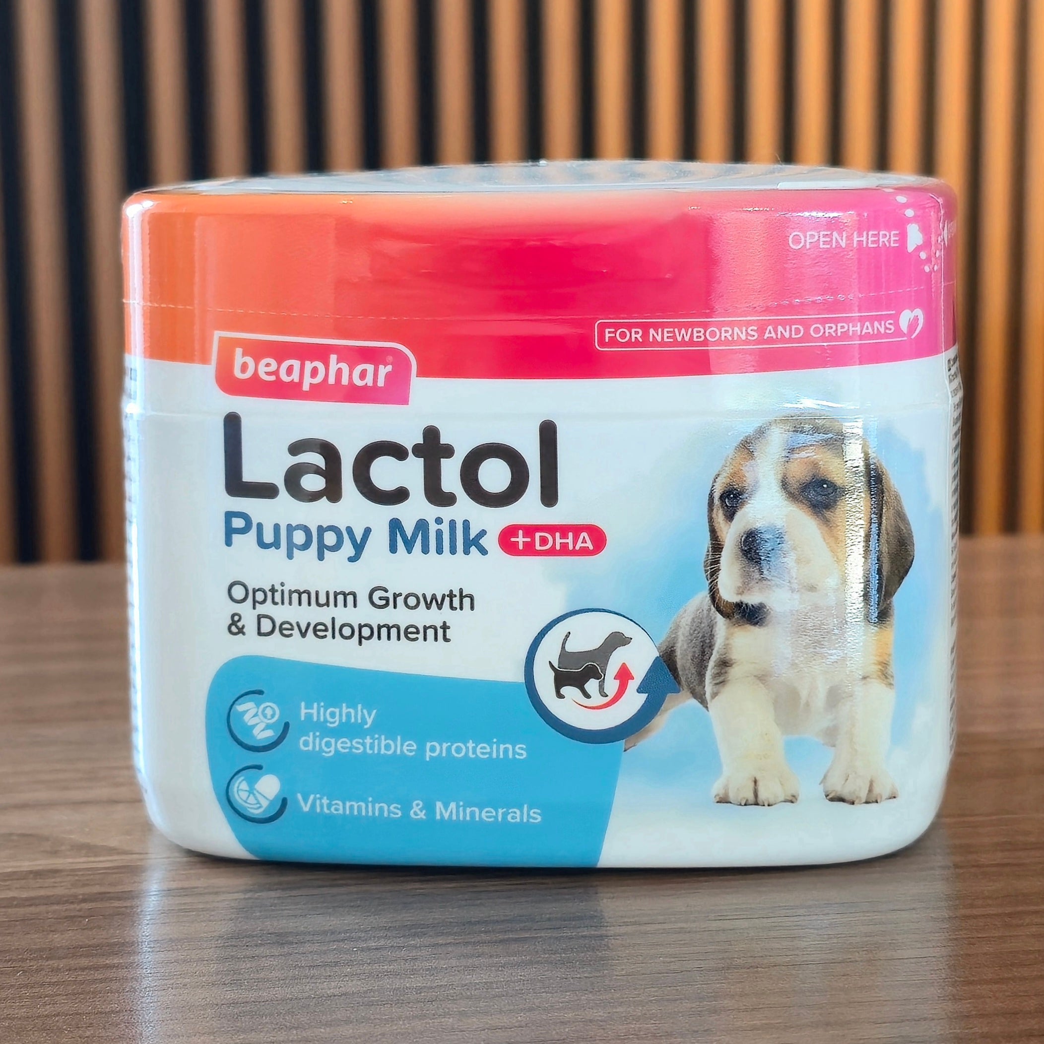 A tub of Beaphar Lactol Puppy Milk +DHA dog formula, featuring a white and blue label with a puppy graphic, set on a wooden table with a slatted wooden background.