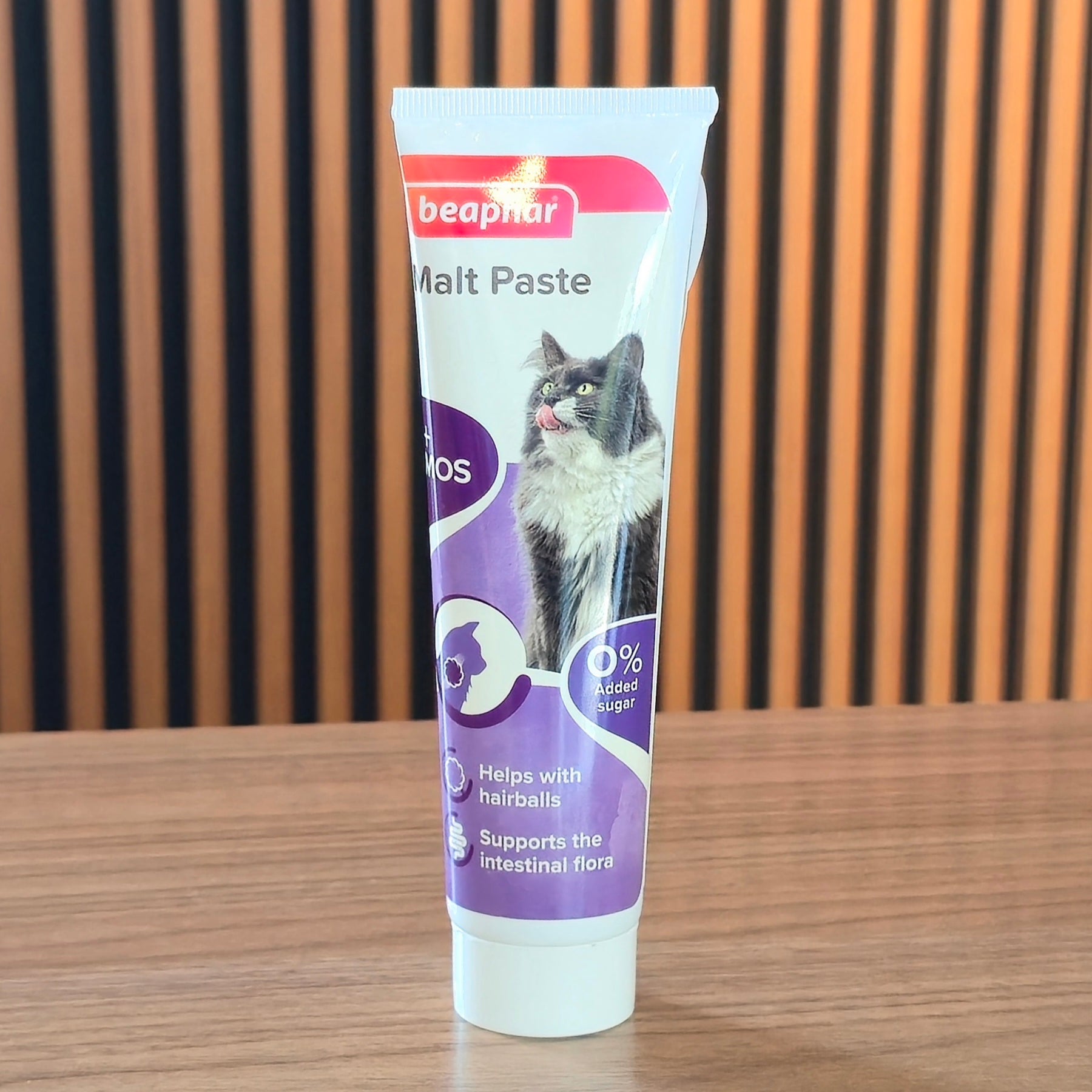 A tube of beaphar Malt Paste cat hairball treatment standing upright on a wooden table, with a slatted wooden wall background.